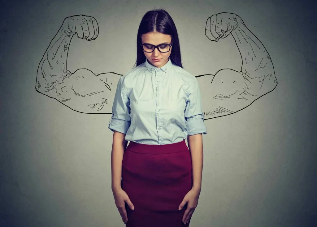 Disrespect destroys self-confidence: a young woman in glasses looks down while strong flexed arms are drawn in chalk on the wall behind her